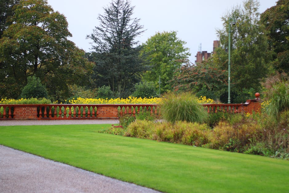 A neatly maintained park border area featuring a well-manicured, lush green lawn in the foreground with even, trimmed grass. Adjacent to the lawn, there is a vibrant garden bed filled with ornamental
